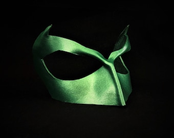 Può includere: Una maschera per gli occhi verde con finitura satinata, caratterizzata da un design curvo e bordi appuntiti. La maschera è posta su uno sfondo nero, evidenziando la sua forma e consistenza. Questo accessorio è adatto per feste in costume o spettacoli teatrali.