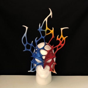 May include: A red, blue, and yellow masquerade mask with a detailed, intricate design. The mask is made of leather and has a delicate, lacy pattern. It is displayed on a white mannequin head.