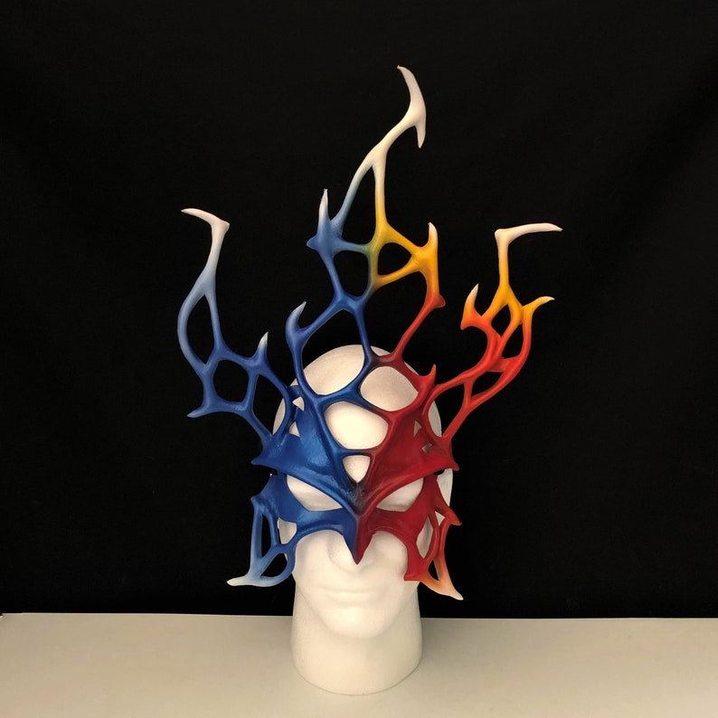 May include: A red, blue, and yellow masquerade mask with a detailed, intricate design. The mask is made of leather and has a delicate, lacy pattern. It is displayed on a white mannequin head.