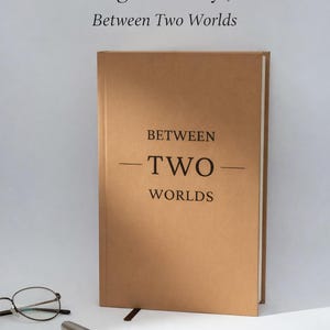 May include: A tan journal with the title "An Immigrant Story Journal Between Two Worlds" in black text. A pair of glasses, a pen, and a fountain pen are on the surface. The journal has a bookmark.