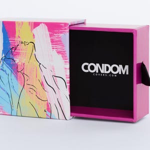 Może przedstawiać: Różowe pudełko z czarnym wnętrzem i białym napisem "CONDOM COVERS.COM". Pudełko jest otwarte, ukazując wnętrze. Na zewnątrz znajduje się kolorowy abstrakcyjny wzór z różowymi, niebieskimi, żółtymi i białymi pociągnięciami pędzla.