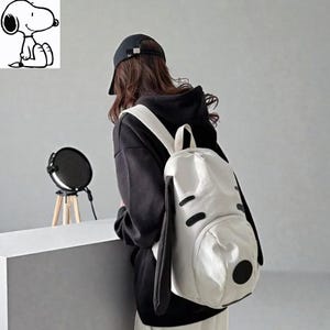 May include: A white backpack shaped like Snoopy's face, featuring black ears, eyes, and a nose. The backpack has a top handle and adjustable straps. The person in the image is wearing a black hoodie and a black cap.