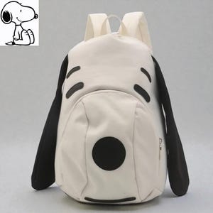 Może przedstawiać: Kremowy plecak z motywem Snoopy'ego. Plecak ma czarne akcenty dla uszu, oczu i nosa, z dużym czarnym kołem dla nosa. Posiada dwa paski na ramiona i górny uchwyt.