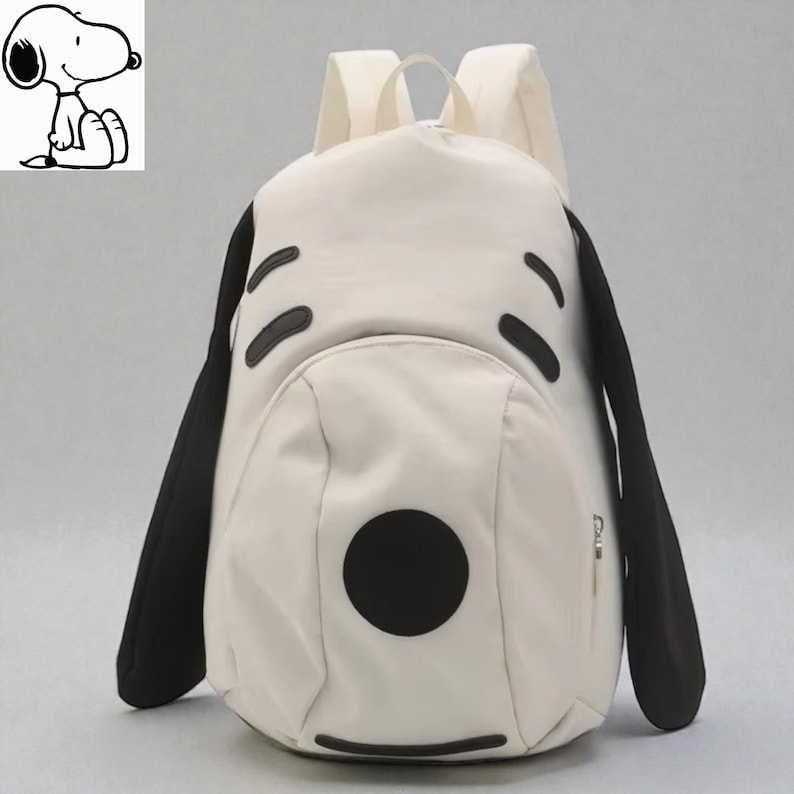 May include: A cream-colored backpack featuring a Snoopy design. The backpack has black accents for the ears, eyes, and nose, with a large black circle for the nose. The backpack has two shoulder straps and a top handle.