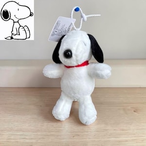 Op de afbeelding: Een witte Snoopy pluche speelgoed met zwarte oren en een rode kraag. Het speelgoed heeft een zwarte neus en staat met uitgestrekte armen. Een klein label is aan de bovenkant van het speelgoed bevestigd.