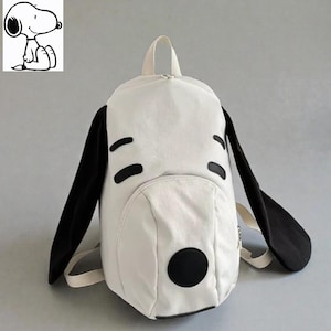 May include: White backpack with black accents, designed to resemble Snoopy's face. Features black ears, eye markings, and a nose. Includes a small front pocket and a top handle. The backpack has adjustable straps.