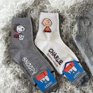 以下が含まれることがあります： ピーナッツのスヌーピーとチャーリー・ブラウンが描かれたクルーソックス2足組。片方はスヌーピーがハートを持っているグレー、もう片方はチャーリー・ブラウンが描かれた白。どちらの靴下にもPeanuts Worldwideのロゴが入っています。