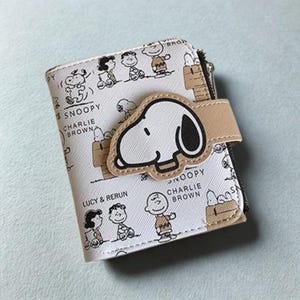 Puede incluir: Una cartera blanca con Snoopy y otros personajes de Peanuts. Tiene una correa beige con un aplique de Snoopy. El diseño incluye el texto "Snoopy" y "Charlie Brown".