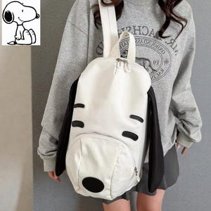 May include: A white backpack with black accents, designed to resemble Snoopy from the Peanuts comic strip. The backpack features black ears, eye markings, and a nose, with a zippered front pocket and a top handle. The bag has a white shoulder strap.