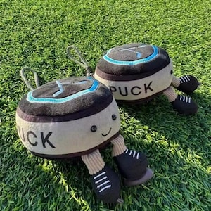 Puede incluir: Dos juguetes de peluche con forma de disco de hockey con caras sonrientes y piernas adjuntas. Los discos son negros y beige con la palabra "PUCK" impresa en el lateral. Cada disco tiene un diseño azul y negro en la parte superior y patines negros.