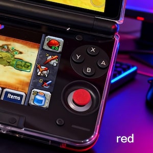 Puede incluir: Primer plano de una consola de juegos portátil negra con una funda protectora transparente. La consola presenta un joystick rojo, botones negros etiquetados X, Y, A y B, y una pantalla que muestra una interfaz de juego. La palabra "red" es visible.