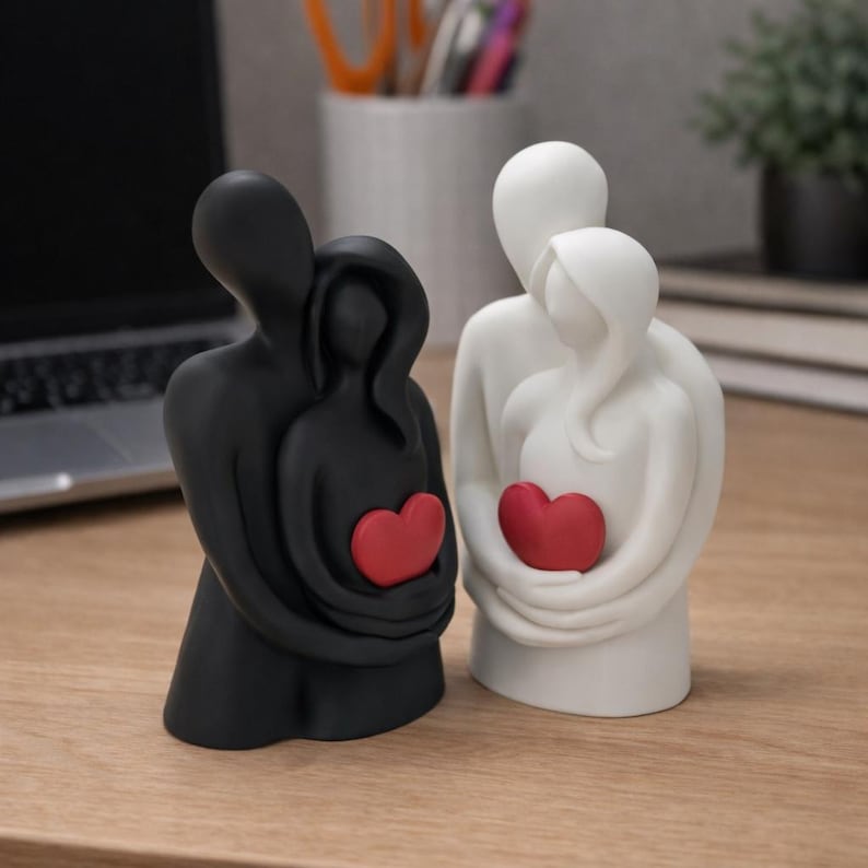 May include: Two ceramic figurines depicting embracing couples, one black and one white. Each figure holds a red heart. The figurines are on a wooden surface, suggesting a theme of love and relationships.