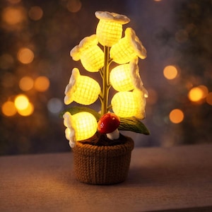May include: A decorative lamp shaped like a flower arrangement. The lamp features yellow, glowing flower-shaped lights with white accents, a red mushroom, and green leaves, all in a brown knitted pot. The background is blurred with warm, out-of-focus lights.