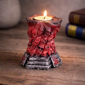 May include: A red and black candle holder with a skull design. The candle holder has a square base and a cylindrical body covered in skulls. A lit tea light candle sits in the top. The candle holder is made of a dark red material.
