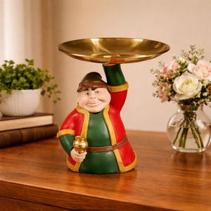 May include: A decorative figurine of a smiling man holding a gold-colored tray above his head. The figurine is dressed in a red coat with yellow trim over a green tunic. He holds a small gold cup in his other hand. The tray is round.