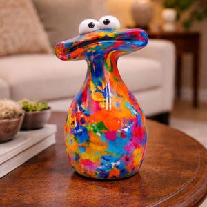 Peut inclure: Un vase en céramique coloré et fantaisiste avec un design abstrait unique. Le vase présente une base bulbeuse et un long col fin, surmonté d'une large tête plate et d'yeux globuleux. La surface est recouverte d'un mélange vibrant de peinture bleue, orange, rose et jaune.