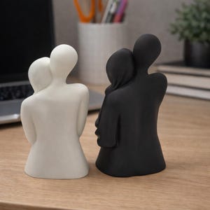 May include: Two minimalist sculptures of embracing figures. One is white, the other is black. The figures are stylized, with smooth, rounded forms. They are positioned on a wooden surface, suggesting a decorative or artistic theme.
