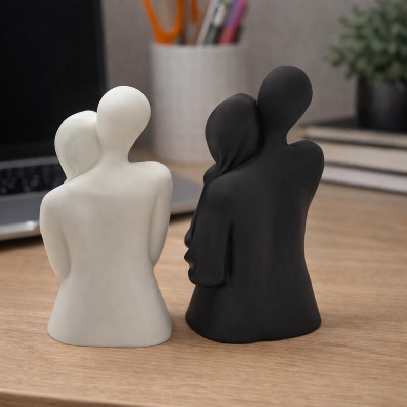 May include: Two minimalist sculptures of embracing figures. One is white, the other is black. The figures are stylized, with smooth, rounded forms. They are positioned on a wooden surface, suggesting a decorative or artistic theme.