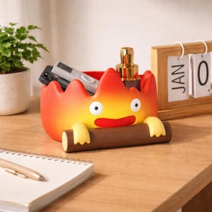 May include: A novelty desk tidy in the shape of a cartoon flame, with red, orange, and yellow hues. It contains small items, including a pen and a calendar. The flame character is holding a brown log. A calendar shows 'JAN 0'.