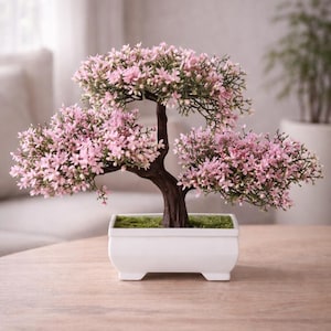 May include: A miniature bonsai tree with pink blossoms and a dark brown trunk sits in a white rectangular pot. The pot rests on a wooden surface. The tree's leaves are a vibrant green.