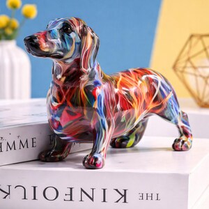 Peut inclure: Figurine de teckel colorée avec un motif de peinture abstrait. La statue de chien est posée sur un livre blanc avec le mot "KINFOLK" imprimé sur le dos. Les couleurs de la figurine comprennent le rouge, le bleu, le jaune et le vert.
