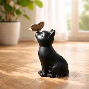 Op de afbeelding: Een zwart kattenbeeldje met een vlinder op zijn neus. De kat zit en kijkt omhoog. De vlinder heeft bruine en beige vleugels. Het beeldje staat op een houten vloer.