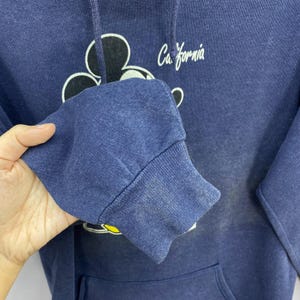 K&ouml;nnte beinhalten: Marineblauer Kapuzenpullover mit wei&szlig;em Mickey Mouse-Design und dem Wort "California" in Schrift. Der Hoodie hat eine Kapuze mit Kordelzug und eine K&auml;ngurutasche. Das B&uuml;ndchen des &Auml;rmels ist sichtbar.