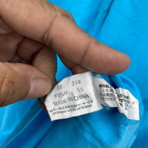 以下が含まれることがあります： 日本語のテキストと「MADE IN CHINA」の文字が入ったタグのクローズアップ。タグには、生地の組成が綿94％、ポリウレタン6％と表示されています。背景は明るい青色の生地です。