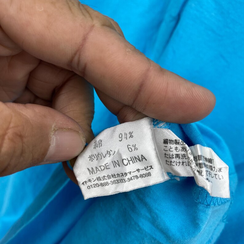 以下が含まれることがあります： 日本語のテキストと「MADE IN CHINA」の文字が入ったタグのクローズアップ。タグには、生地の組成が綿94％、ポリウレタン6％と表示されています。背景は明るい青色の生地です。