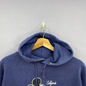 K&ouml;nnte beinhalten: Marineblauer Kapuzenpullover mit Kordelzug und einer Grafik von Mickey Mouse und dem Wort "California" in Schreibschrift. Der Pullover h&auml;ngt an einem Holzb&uuml;gel.