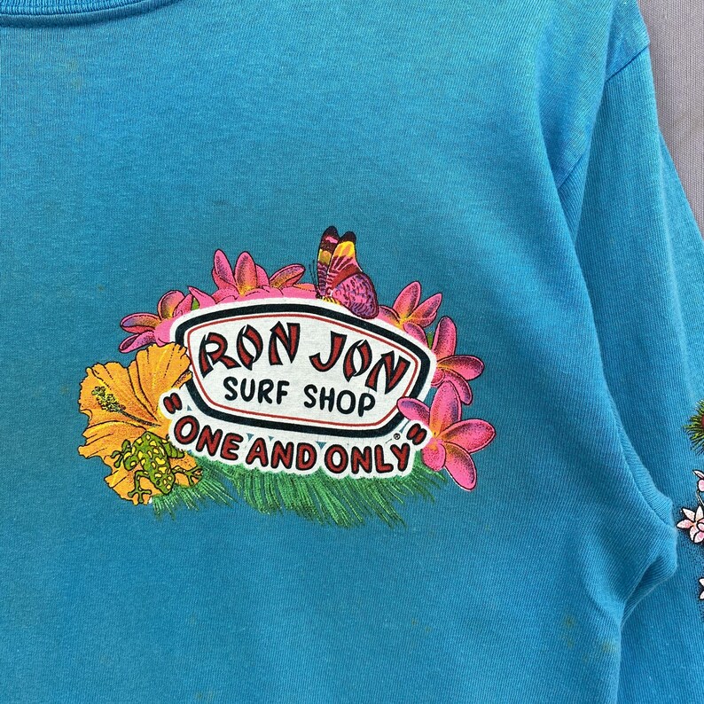 K&ouml;nnte beinhalten: Ein hellt&uuml;rkisfarbenes Langarmshirt mit einem Grafikdruck, der einen Schmetterling, rosa Frangipani-Bl&uuml;ten, einen gelben Hibiskus und eine gr&uuml;ne Eidechse zeigt. Der Text RON JON SURF SHOP und ONE AND ONLY steht in einem wei&szlig;en Oval.