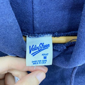 K&ouml;nnte beinhalten: Nahaufnahme eines marineblauen Kapuzenpullovers mit einem wei&szlig;en Etikett. Auf dem Etikett steht "Velva Sheen" in blauer Schrift, mit "ADULT M" und "OVER FOR CARE MADE IN U.S.A." darunter. Der Pullover ist aus einem weichen, strukturierten Stoff.