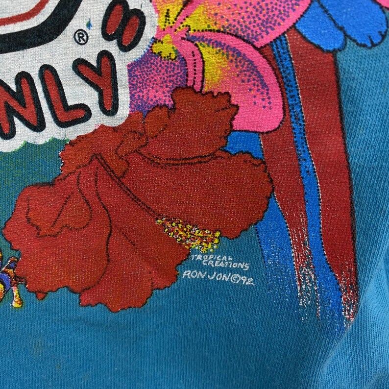 K&ouml;nnte beinhalten: Ein t&uuml;rkisfarbener Stoff mit einem lebendigen Grafikdruck einer gro&szlig;en roten Hibiskusbl&uuml;te, rosa tropischen Bl&uuml;tenbl&auml;ttern und blauen Akzenten. Der Text lautet TROPICAL CREATIONS RON JON 92, wobei teilweise schwarze und rote Buchstaben zu sehen sind.