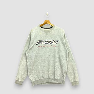 以下が含まれることがあります： 胸に大胆で様式化されたフォントで「FUBU」という文字が入ったライトグレーのスウェットシャツ。ブランド名のすぐ下に、「THE COLLECTION」というテキストが小さな赤いフォントで表示されています。スウェットシャツは木製のハンガーに吊るされています。