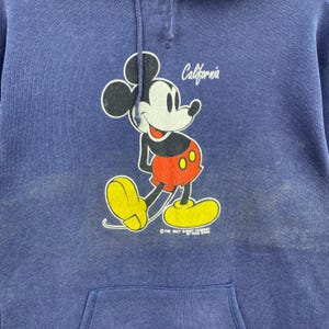 K&ouml;nnte beinhalten: Blauer Kapuzenpullover mit einem Mickey Mouse-Motiv. Mickey ist in Bewegung dargestellt und tr&auml;gt rote Shorts und gelbe Schuhe. Das Wort "California" steht &uuml;ber der Figur. Unten steht "The Walt Disney Company by Veda Brien".