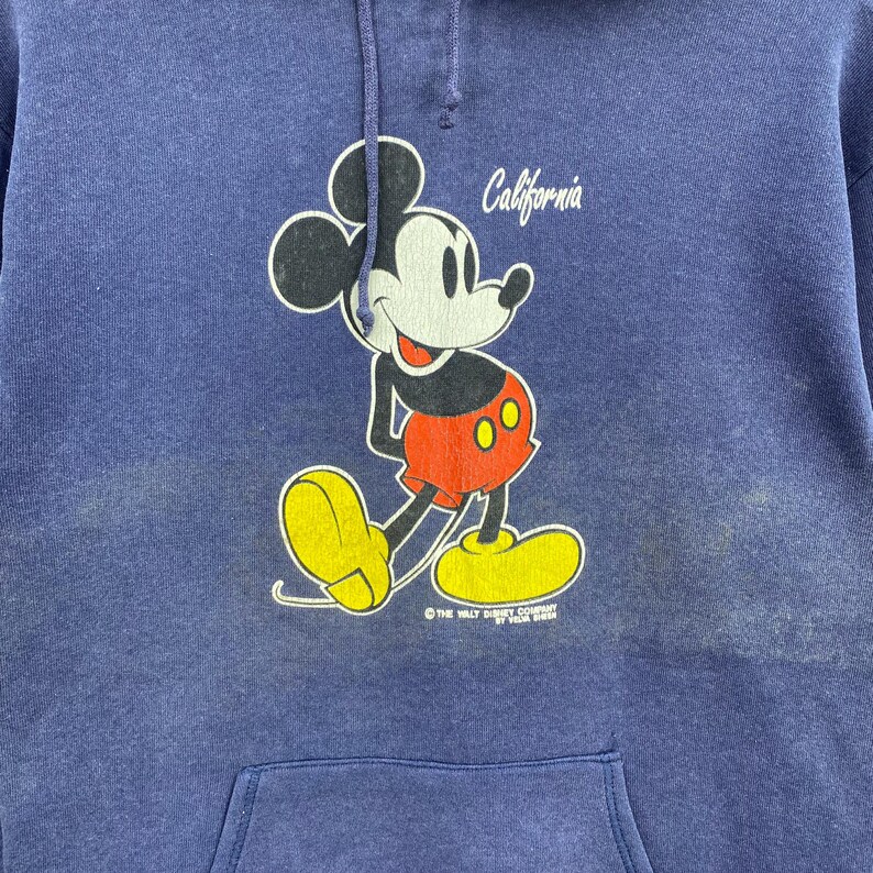 K&ouml;nnte beinhalten: Blauer Kapuzenpullover mit einem Mickey Mouse-Motiv. Mickey ist in Bewegung dargestellt und tr&auml;gt rote Shorts und gelbe Schuhe. Das Wort "California" steht &uuml;ber der Figur. Unten steht "The Walt Disney Company by Veda Brien".