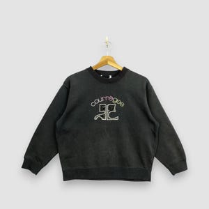 May include: A black crew neck sweatshirt with the word "courr&egrave;ges" in colorful embroidery above a stylized design. The sweatshirt is hanging on a wooden hanger against a light background.