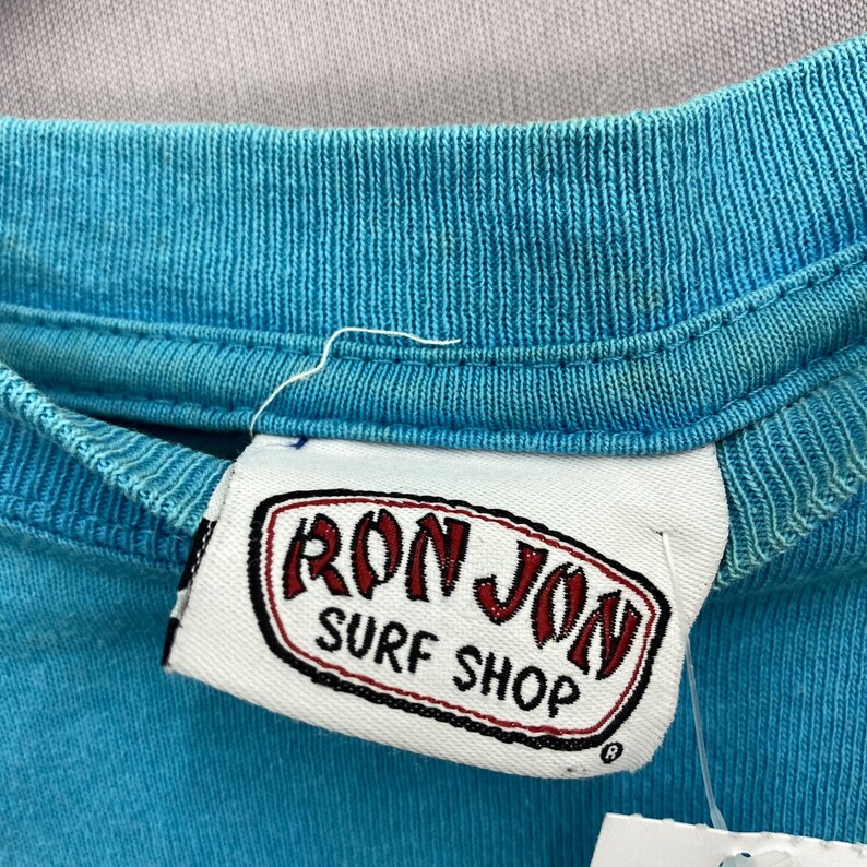 K&ouml;nnte beinhalten: Ein hellt&uuml;rkisfarbenes T-Shirt mit einem wei&szlig;en Stoffetikett am Kragen, das ein rot-schwarzes Logo mit der Aufschrift RON JON SURF SHOP zeigt. Das Etikett ist in den inneren Kragen des Kleidungsst&uuml;cks eingen&auml;ht, das eine gerippte Struktur aufweist.