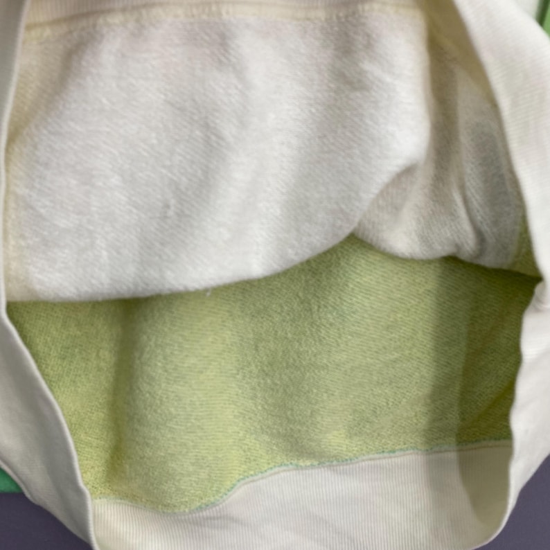 Puede incluir: Primer plano del interior de una sudadera blanca y verde claro. El interior presenta una tela suave y texturizada en blanco y verde claro. El dise&ntilde;o de la sudadera incluye un dobladillo y pu&ntilde;os acanalados, con una construcci&oacute;n en capas.