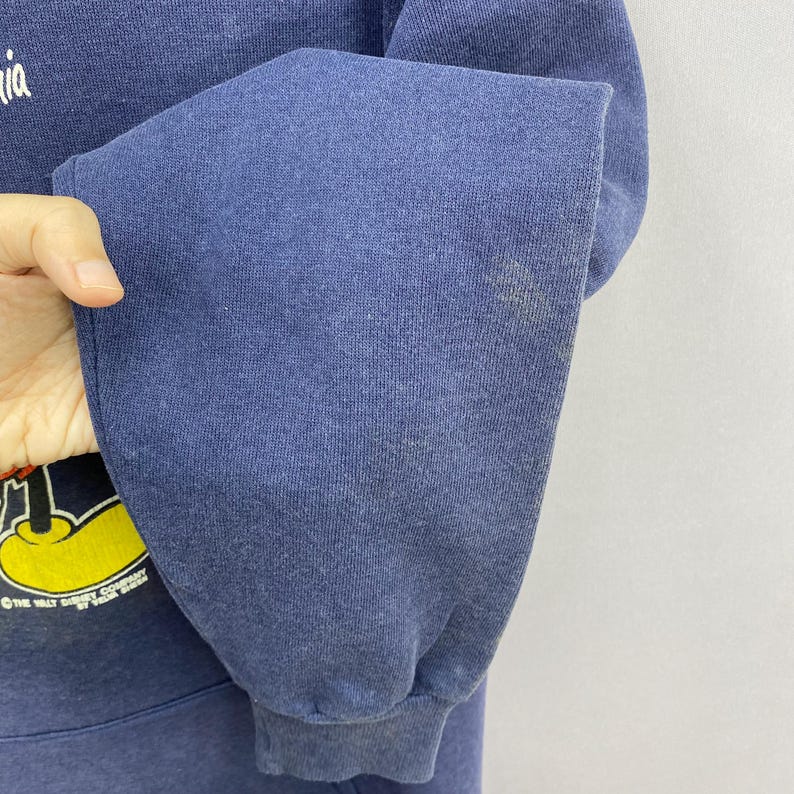 K&ouml;nnte beinhalten: Ein marineblaues Sweatshirt mit einer Mickey Mouse-Grafik. Der &Auml;rmel weist einen Fleck auf. Das Logo der Walt Disney Company ist teilweise sichtbar. Das Sweatshirt besteht aus einem weichen, bequemen Material.