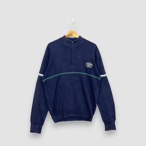 May include: Navy blue zip-up sweater with a green and white stripe across the chest and on the left sleeve. The sweater has a small logo on the chest. The sweater is hanging on a wooden hanger.