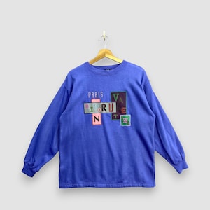 Peut inclure: Un sweat-shirt bleu à manches longues avec un imprimé graphique sur la poitrine comportant le texte PARIS et VALENTINO disposé dans des blocs rectangulaires colorés. Le motif présente des lettres de polices et couleurs variées.