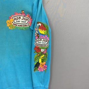 K&ouml;nnte beinhalten: Ein helles t&uuml;rkisfarbenes Langarmshirt mit einer Grafik auf der Brust und dem &Auml;rmel. Die Brustgrafik zeigt ein Ron Jon Surf Shop One and Only Logo mit Blumen und einem Schmetterling. Der &Auml;rmel zeigt einen Papagei, einen Tukan und Blumen.