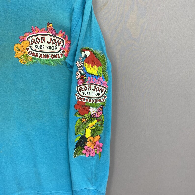K&ouml;nnte beinhalten: Ein helles t&uuml;rkisfarbenes Langarmshirt mit einer Grafik auf der Brust und dem &Auml;rmel. Die Brustgrafik zeigt ein Ron Jon Surf Shop One and Only Logo mit Blumen und einem Schmetterling. Der &Auml;rmel zeigt einen Papagei, einen Tukan und Blumen.