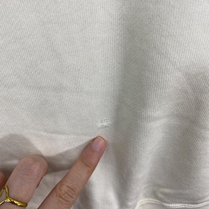 Puede incluir: Primer plano de una tela de sudadera de color crema con un peque&ntilde;o agujero. La tela tiene una textura acanalada. Un dedo con un anillo de oro se&ntilde;ala el da&ntilde;o.