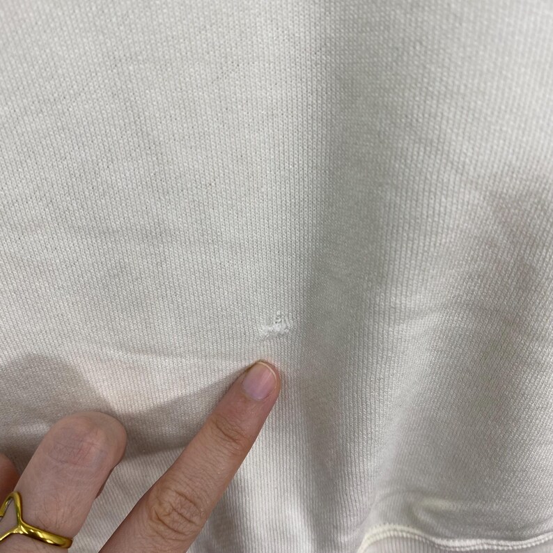 Puede incluir: Primer plano de una tela de sudadera de color crema con un peque&ntilde;o agujero. La tela tiene una textura acanalada. Un dedo con un anillo de oro se&ntilde;ala el da&ntilde;o.