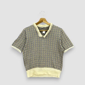 May include: A short-sleeved, collared shirt with a checkered pattern in shades of brown, navy, and olive green. The collar, cuffs, and hem are a light cream color. The shirt is hanging on a wooden hanger.