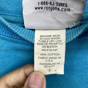 K&ouml;nnte beinhalten: Ein leuchtend blaues Baumwoll-T-Shirt mit einem wei&szlig;en Pflegeetikett auf der Innenseite. Das Etikett zeigt den Text 1-888-RJ-SURFS, www.ronjons.com, MACHINE WASH IN COLD WATER, TUMBLE DRY LOW, REMOVE PROMPTLY, NO BLEACH, RN #101747, 100% COTTON, MADE IN U.S.A., ASSEMBLED IN MEXICO und die Gr&ouml;&szlig;e S.