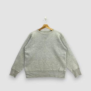 May include: A heather gray long-sleeve crewneck sweatshirt featuring a ribbed collar, cuffs, and hem. The garment includes a triangular stitched V-insert detail at the center of the neckline and is displayed on a wooden hanger against a plain background.