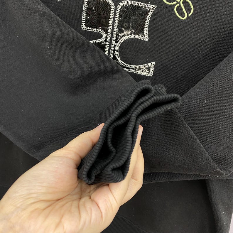May include: Black sweatshirt with a sequined design and yellow accents. The design appears to be a stylized logo or emblem. The sleeve cuff is ribbed. The sweatshirt is made of a soft material.